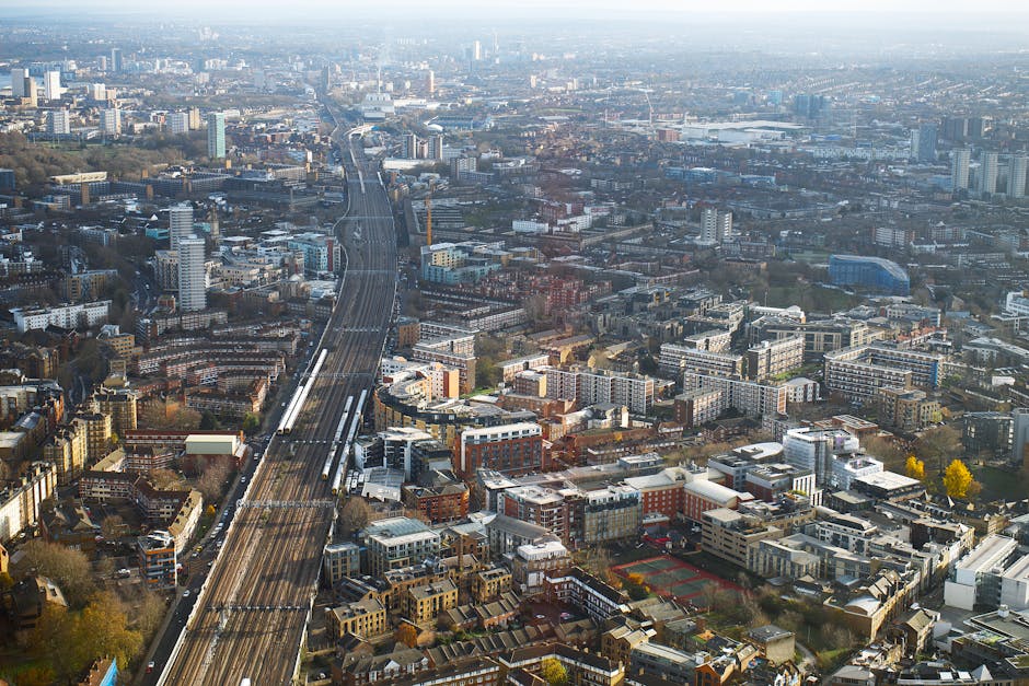 An aerial view of the cityscape in Elephant and Castle, showing a major railway line running through the densely built urban area with multiple modern and traditional residential and commercial buildings. In the foreground, there are several multi-story apartment buildings, some with flat roofs and balconies, alongside smaller houses, with a few trees scattered among them. The railway tracks are visible, with trains stationary or moving along the lines, and adjacent to the tracks is a loading area with a van, partially visible, used for home relocation or furniture transport. The environment appears well-lit with natural daylight, and the overall scene captures the busy, built-up nature of the area, which aligns with the logistical processes of house removals and packing services provided by companies like [COMPANY_NAME], operating in the region. The backdrop extends into the distance with more high-rise buildings, roads, and urban infrastructure, illustrating the extensive scope of moving services in the area around Heygate Estate.