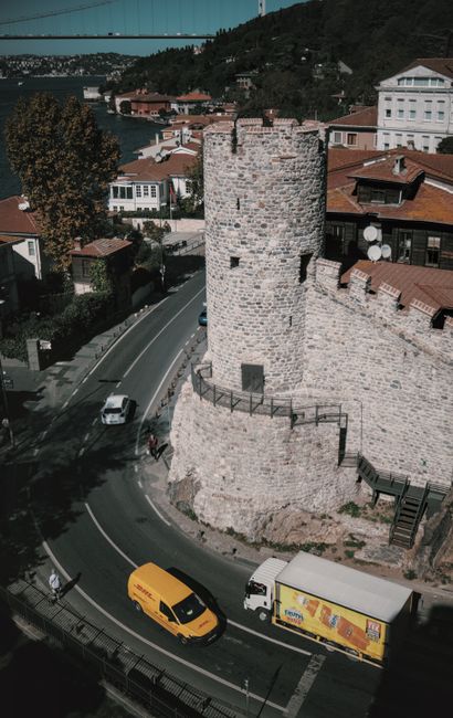 A tall, cylindrical stone tower with a small, rectangular door at its base and a metal railing along its top, situated on a curved street in an urban area. Surrounding the tower are residential buildings with red-tiled roofs, some with satellite dishes, and trees with autumn foliage. Cars, including a yellow van and a white delivery truck with a colorful advertisement, are parked or moving along the street. The scene is captured during daylight with shadows cast by the buildings and vehicles. The background features a body of water, a bridge, and hills with more buildings. This image depicts a typical street scene near historic architecture, relevant to house removals and moving logistics involving residential and commercial premises in an urban setting, as managed by [COMPANY_NAME].