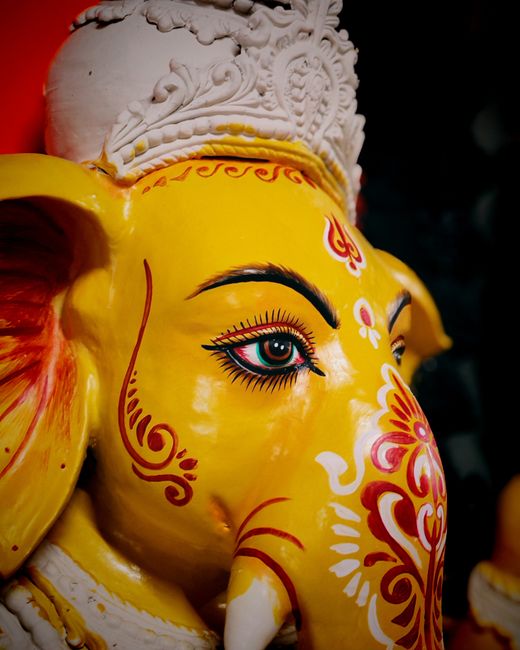 A close-up photograph of a decorative statue of the Hindu deity Ganesha, featuring a yellow face with detailed painted designs in red, white, and black. The statue has large, expressive eyes with pain