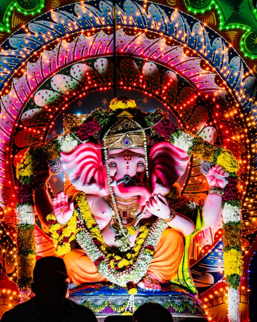 A close-up photograph of a decorative statue of the Hindu deity Ganesha, featuring a yellow face with detailed painted designs in red, white, and black. The statue has large, expressive eyes with pain