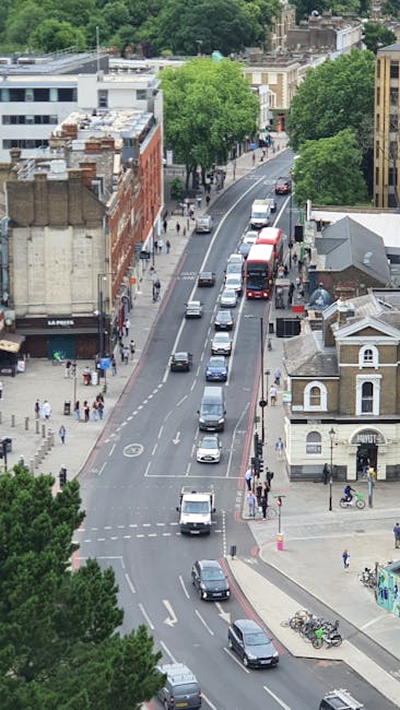 An aerial view of a busy urban street with multiple lanes of traffic, including cars, vans, and a double-decker bus, moving through a city environment with buildings, trees, and pedestrians along the sidewalks. The street features parked bicycles near the curb and small shopfronts, some with awnings. The scene includes a mixture of modern and historic architecture, with green trees lining the road providing a contrast to the concrete structures. The lighting suggests daytime with clear visibility. This setting reflects typical city traffic flow and pedestrian activity, commonly encountered during residential or commercial relocation processes, where belongings may be transported via nearby loading zones or delivery areas, and moving companies such as removalcompanyelephantandcastle.co.uk might be involved in home relocation or furniture transport services.