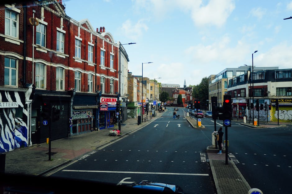 Photograph of a busy urban street scene captured during daytime, viewed from inside a vehicle. The street is lined with a mix of older red-brick buildings with large windows and more modern structures, featuring shops and commercial premises at ground level. On the left side, there are shops with storefronts, some with closed metal shutters, and signage for local businesses. The right side includes a building with graffiti art on its wall and a shop with a yellow sign. The road has multiple lanes with white directional arrows, and traffic lights display red signals for vehicles waiting at the intersection. A few pedestrians are visible on the sidewalks, with some standing near bus stops or crossing the street. The area appears to be part of a neighbourhood undergoing a home relocation or moving process, with the scene suggesting a typical street where removals might load or unload furniture and boxes. The sky is partly cloudy with patches of blue, and streetlights are evenly spaced along the pavement, contributing to a clear, well-lit environment appropriate for house removals and transportation services.